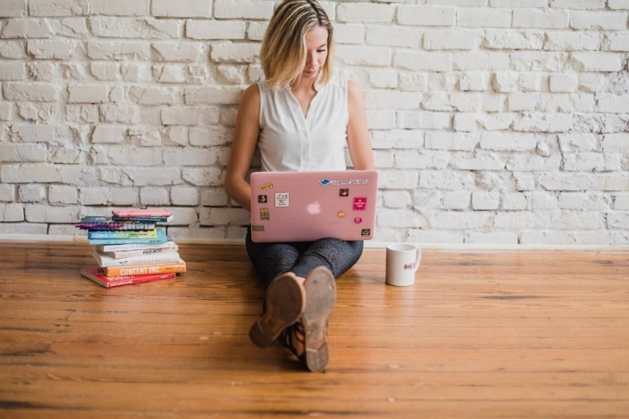 Stockfoto: Zu sehen ist eine Frau, die mit Laptop auf dem Boden sitzt. Neben ihr ist eine Tasse und ein Bücherstapel zu sehen. 